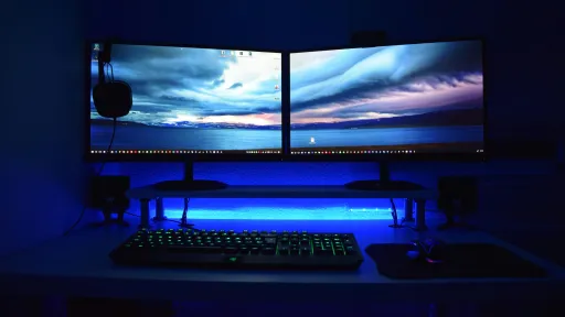 Dual computer monitors with a scenic view, backlit in blue, with a keyboard and headset.